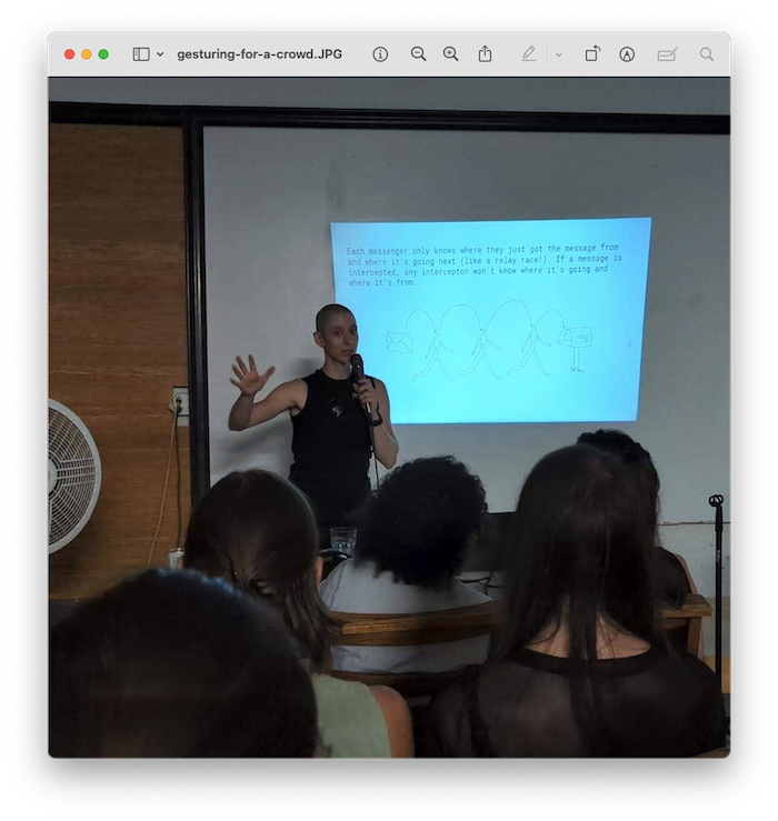 a screenshot titled 'gesturing-for-a-crowd.JPG' that shows a photograph of Lizz holding a microphone and standing in front of a small crowd. Behind Lizz is a presentation slide with text and a hand-drawn diagram.